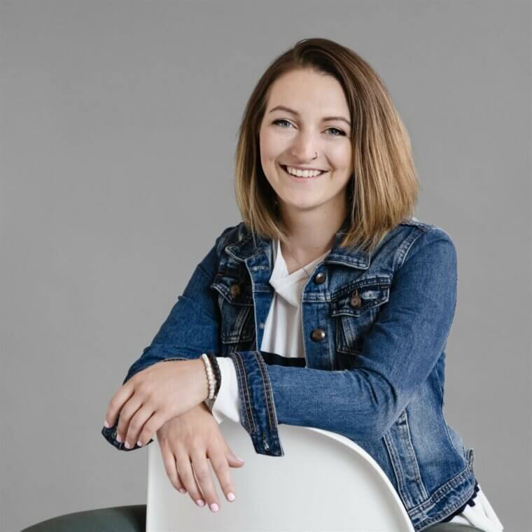 A smiling woman in a denim jacket sitting on a white chair.